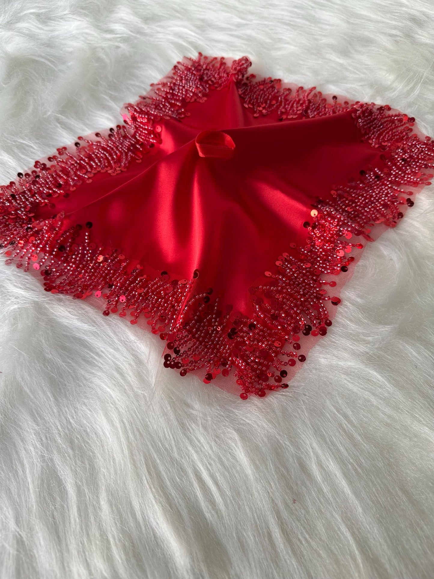 A red satin pillow with a beaded fringe border. The pillow is square shaped and has a hole in the center.