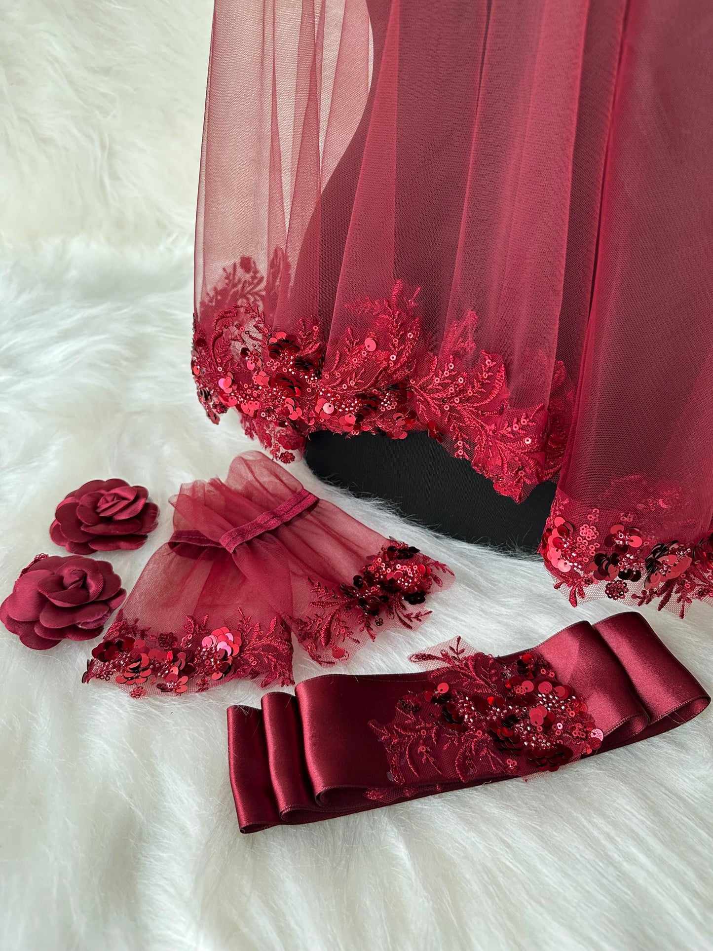 A burgundy bridal veil with lace and sequins, a pair of burgundy lace gloves with sequins, a burgundy satin sash with lace and sequins, and two burgundy fabric flowers.