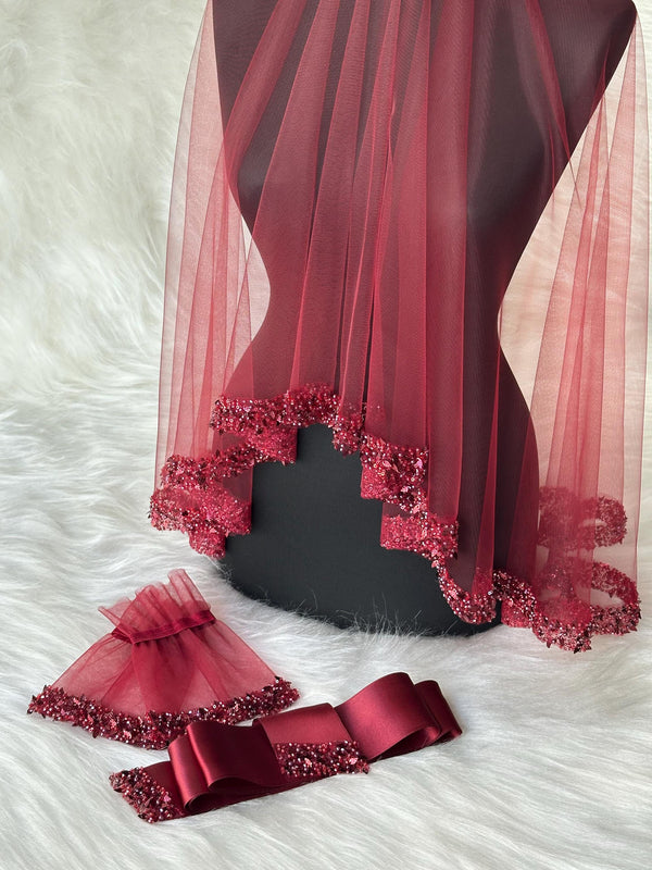 A burgundy tulle veil with a beaded trim. The veil is displayed on a mannequin. There is also a burgundy satin sash and a pair of burgundy tulle wrist cuffs with beaded trim.
