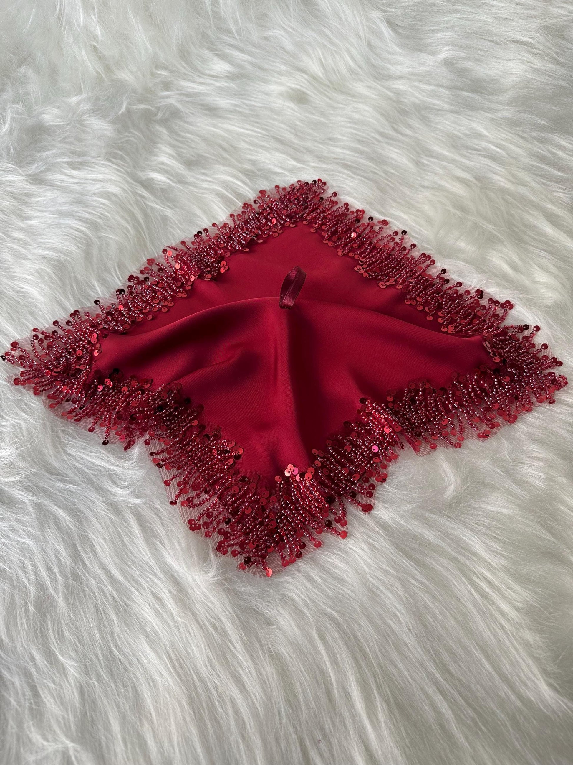 A red square shaped fabric with a beaded fringe around the edges. The fabric is made of a soft, silky material and has a subtle sheen.