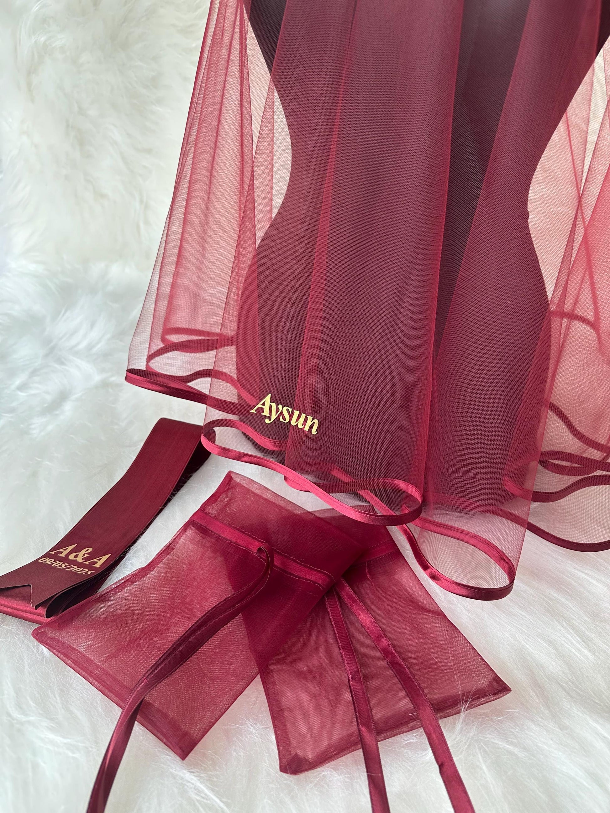 A burgundy tulle veil with a satin trim and the word 'Aysun' embroidered in gold. The veil is displayed on a mannequin. Two burgundy satin drawstring bags with the words 'A&A' and the date '08.08.2023' are also shown.