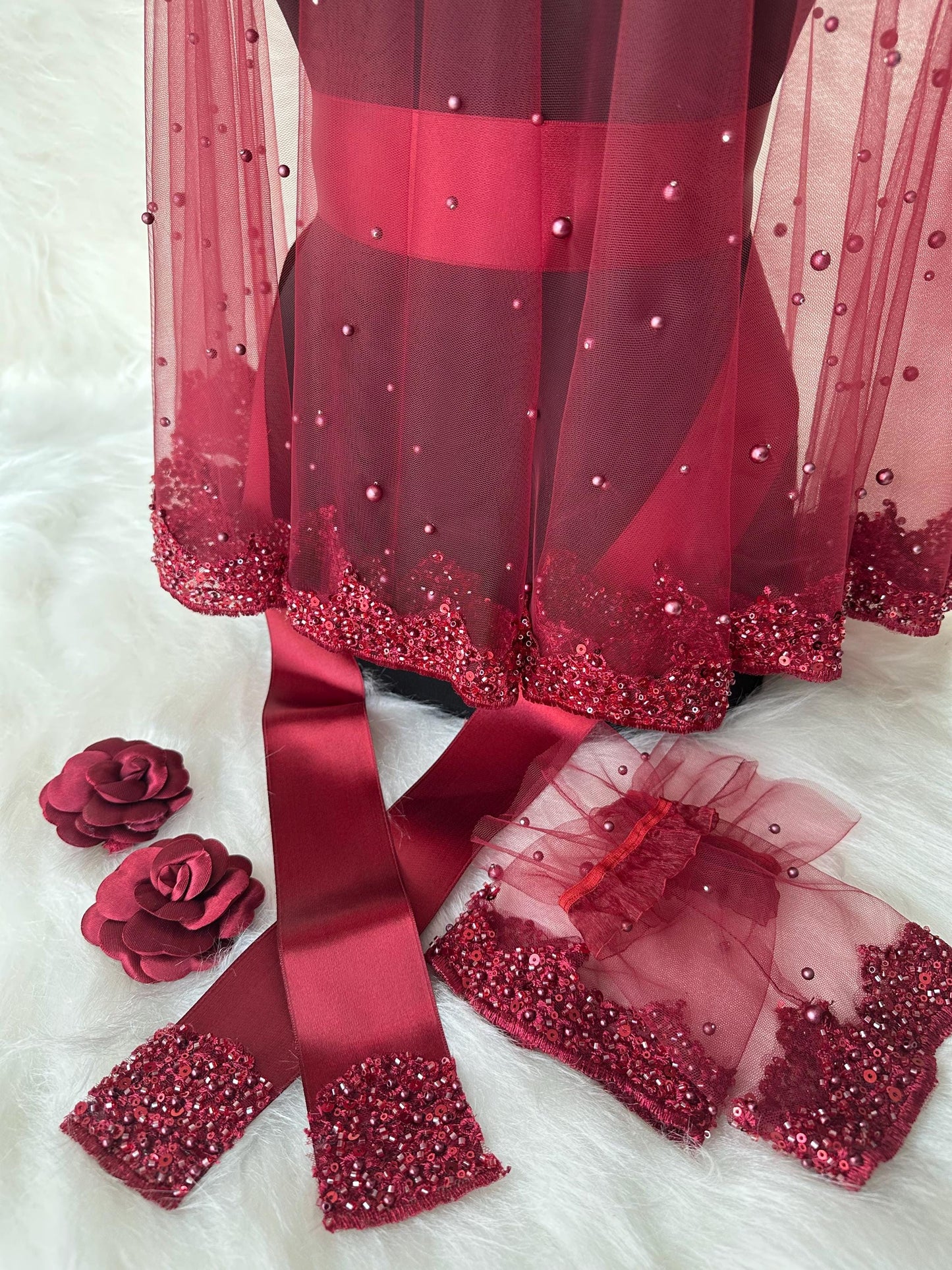 A red  bridal shawl with a beaded trim, a matching burgundy satin ribbon, and two burgundy fabric rose embellishments.