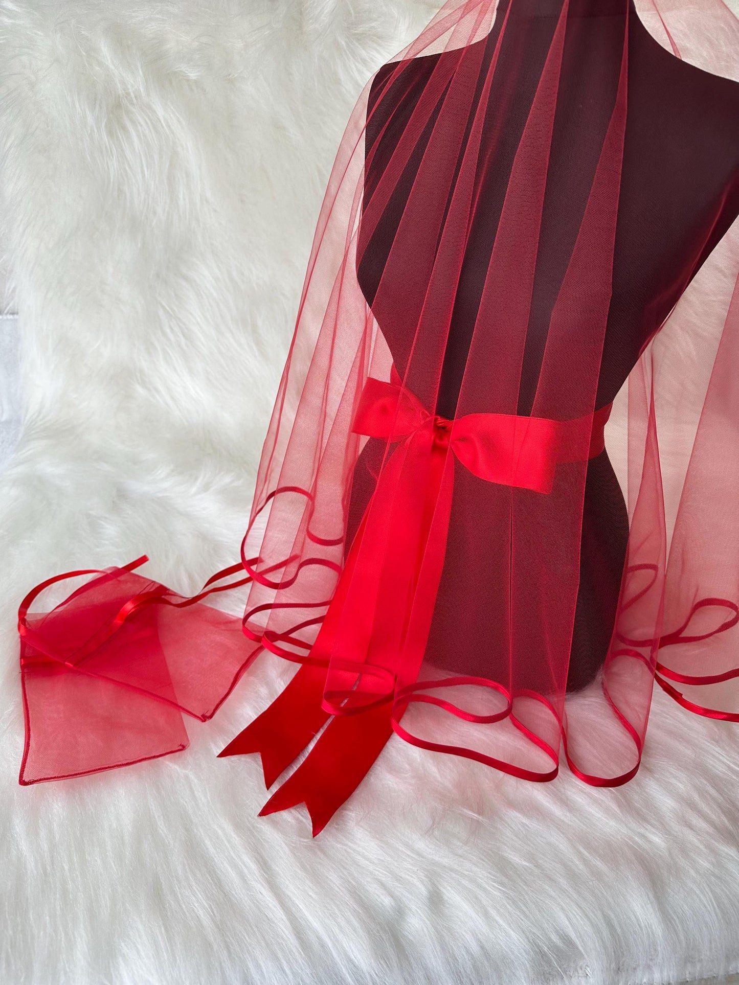 A red tulle bridal veil with a red satin ribbon tied around the waist. The veil has a scalloped edge and the ribbon has a bow in the back.