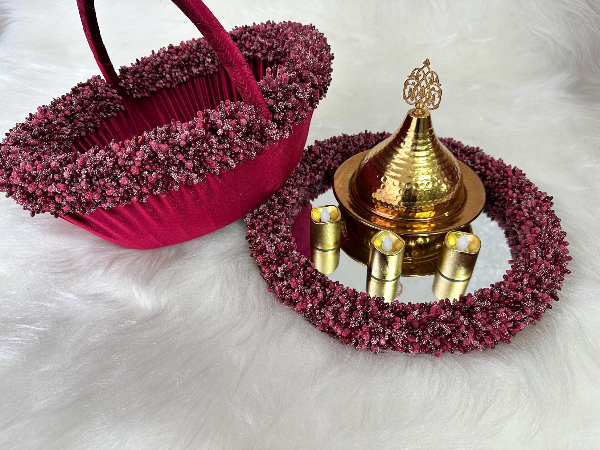 A gold decorative tray with a gold lid and three small candles. The tray is surrounded by a burgundy beaded trim. A burgundy velvet basket with a beaded trim is also in the image.