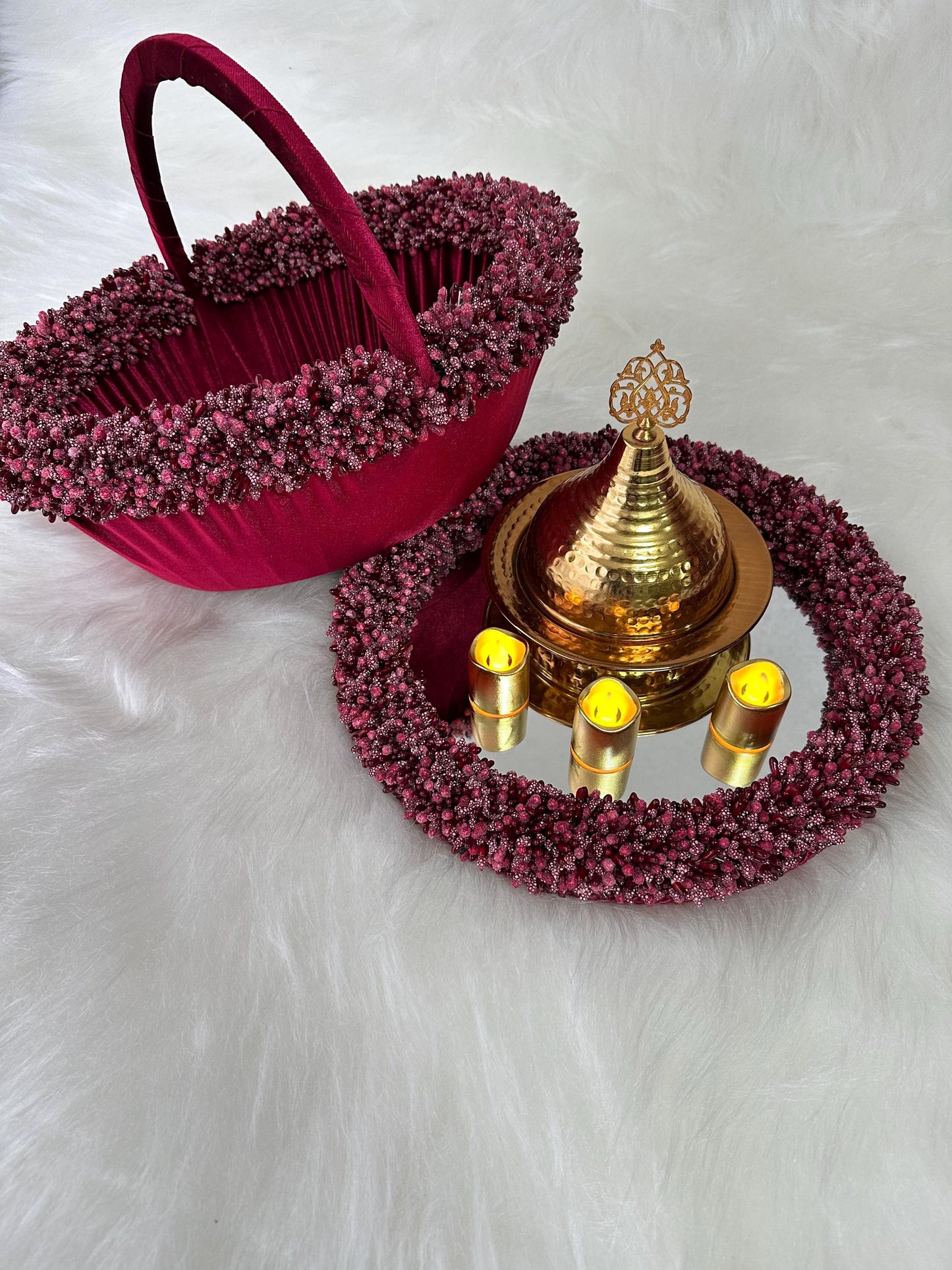A red velvet basket with a gold handle and a gold decorative tray with a gold lid. The basket and tray are surrounded by a pink beaded trim. The tray has four small candles on a mirrored surface.