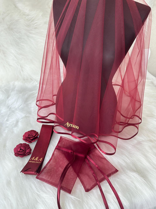 A burgundy tulle veil with a satin ribbon trim and the name 'Aysun' embroidered in gold. The veil is accompanied by two burgundy satin ribbon sashes with the text 'A&A 08.03.2023' and two burgundy tulle pouches with satin ribbon ties.