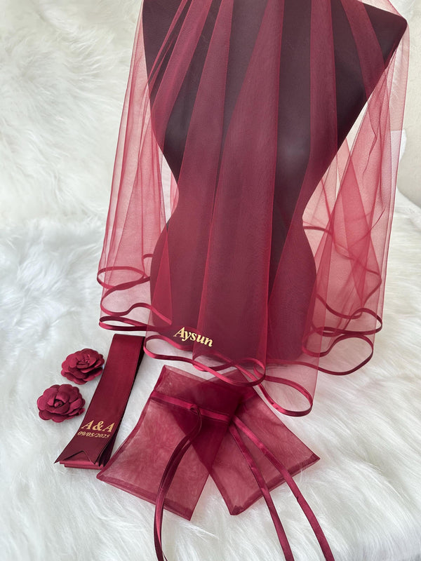 A burgundy tulle veil with a satin ribbon trim and the name 'Aysun' embroidered in gold. The veil is accompanied by two burgundy satin ribbon sashes with the text 'A&A 08.03.2023' and two burgundy tulle pouches with satin ribbon ties.