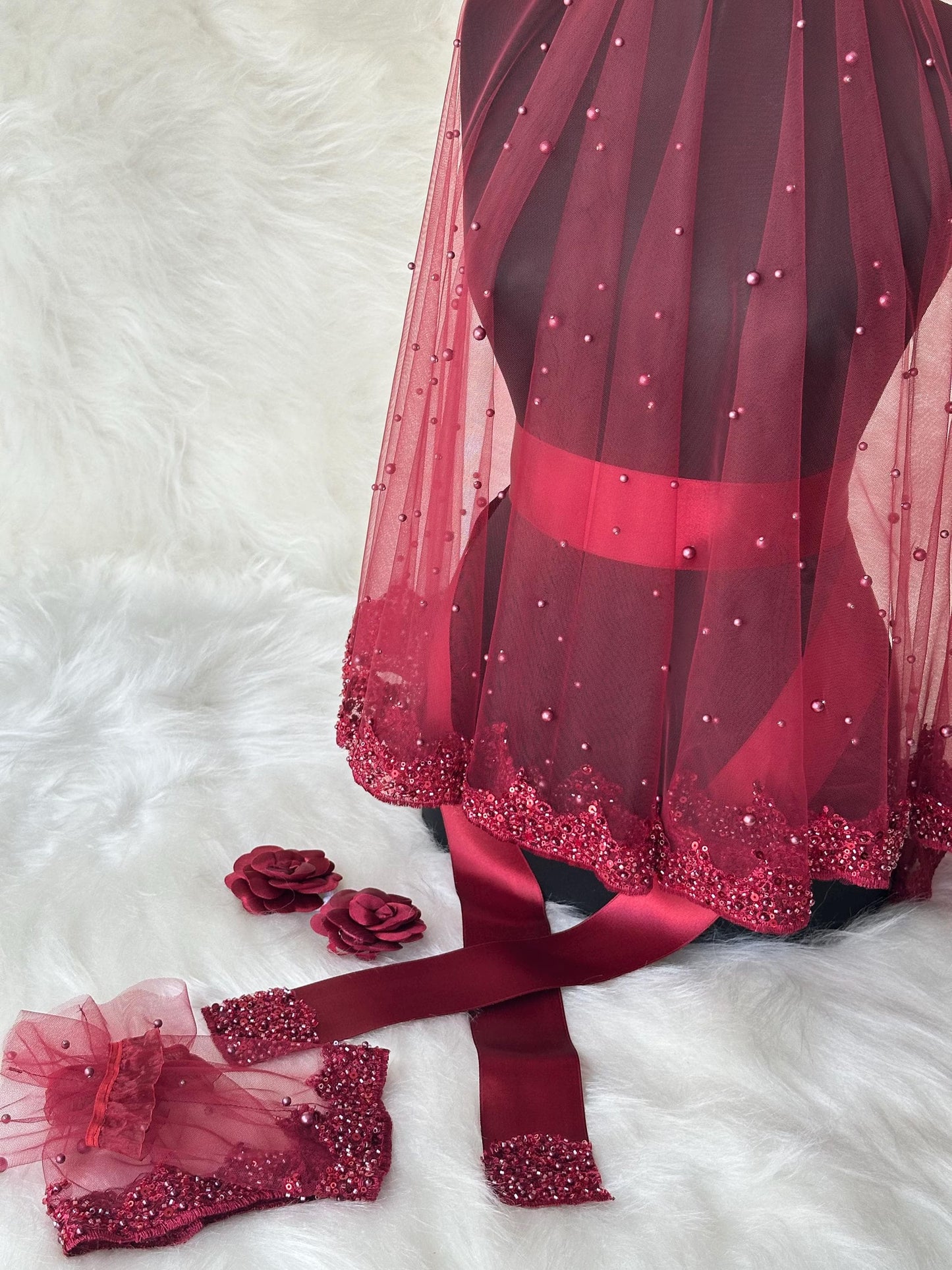 A burgundy bridal veil with pearl embellishments and a matching burgundy satin ribbon. The veil is made of tulle and has a scalloped edge. The ribbon is tied around the waist and has a bow at the back. There are also two burgundy fabric rose embellishments.