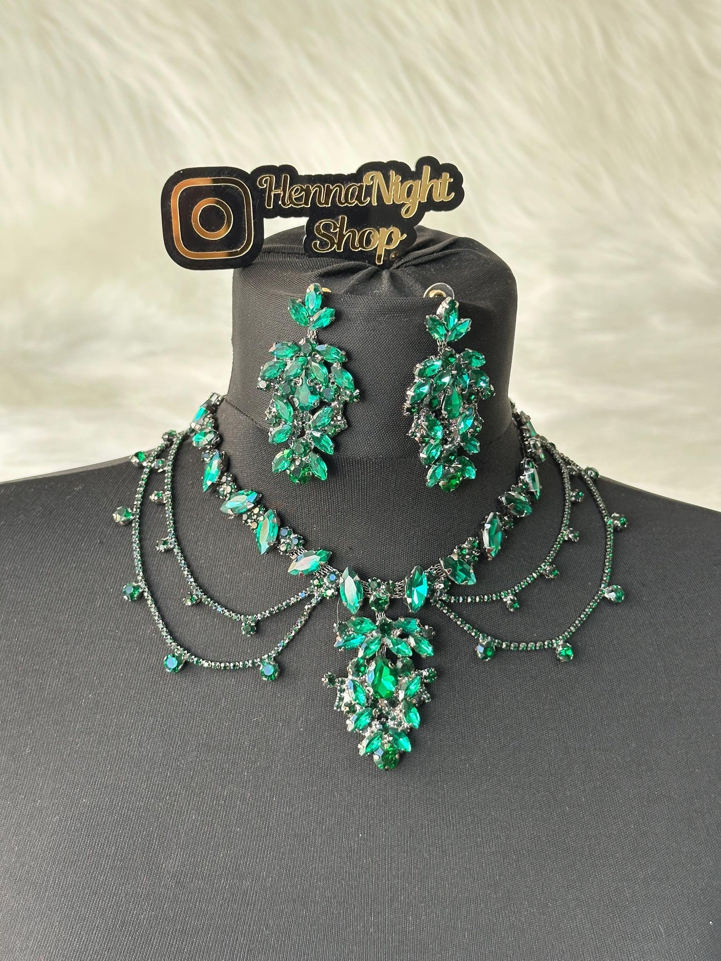 A green crystal necklace and earrings set. The necklace has three strands of small crystals and a large pendant with a cluster of larger crystals. The earrings are long and dangle with a cluster of crystals at the bottom. The jewelry is displayed on a black velvet background. The image also includes a logo that says 'Henna Night Shop'.