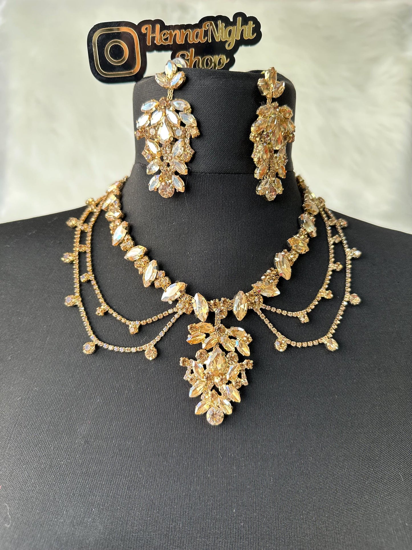 A gold necklace and earrings set with clear and champagne-colored rhinestones. The necklace has a layered design with a large pendant in the center. The earrings are long and dangle. The set is displayed on a black mannequin. The text 'HennaNight Shop' is visible in the top center of the image.