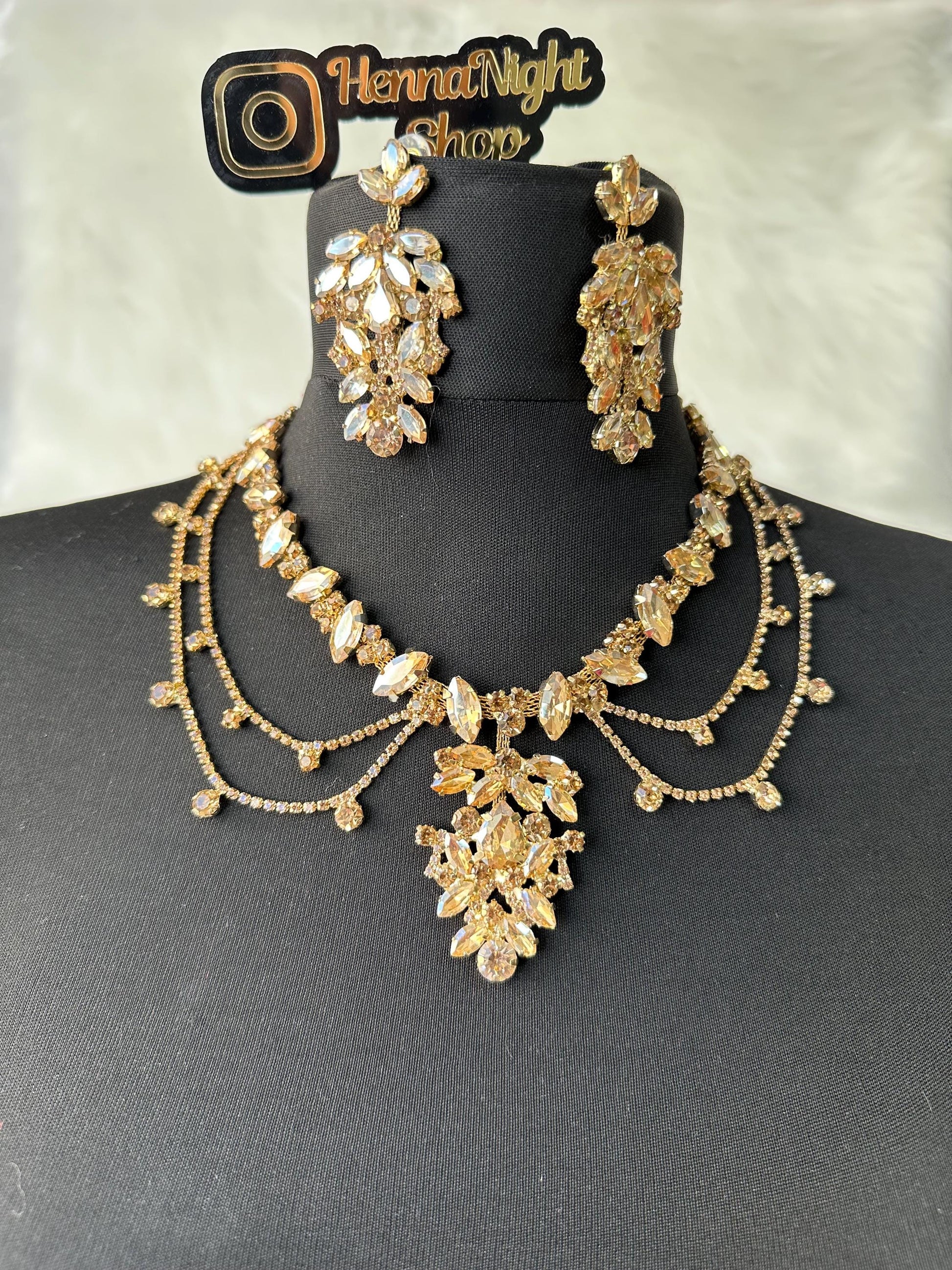 A gold necklace and earrings set with clear and champagne-colored rhinestones. The necklace has a layered design with a large pendant in the center. The earrings are long and dangle. The set is displayed on a black mannequin. The text 'HennaNight Shop' is visible in the top center of the image.