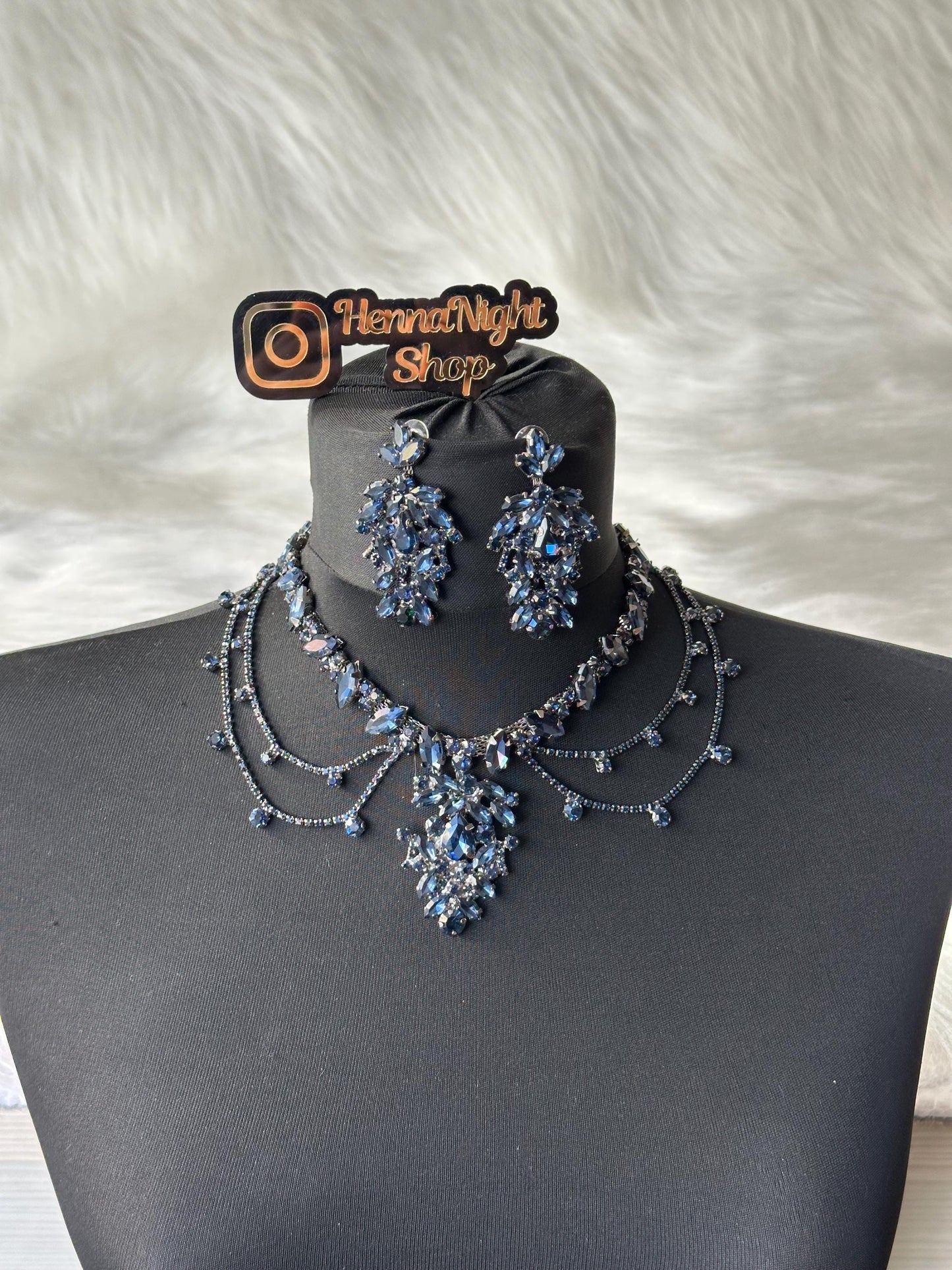 A black choker necklace with a large blue crystal pendant. The necklace has three strands of smaller crystals. Two matching blue crystal earrings are also shown. The image includes the text 'HennaNight Shop'.