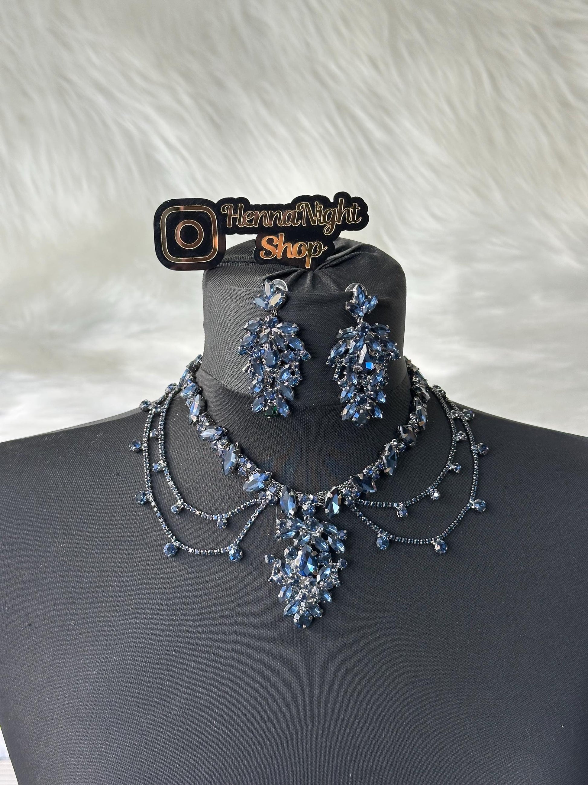 A black necklace with a large blue crystal pendant and matching earrings. The necklace has three strands of small black crystals. The earrings are also made of blue crystals. The image includes a logo that says 'Henna Night Shop'.