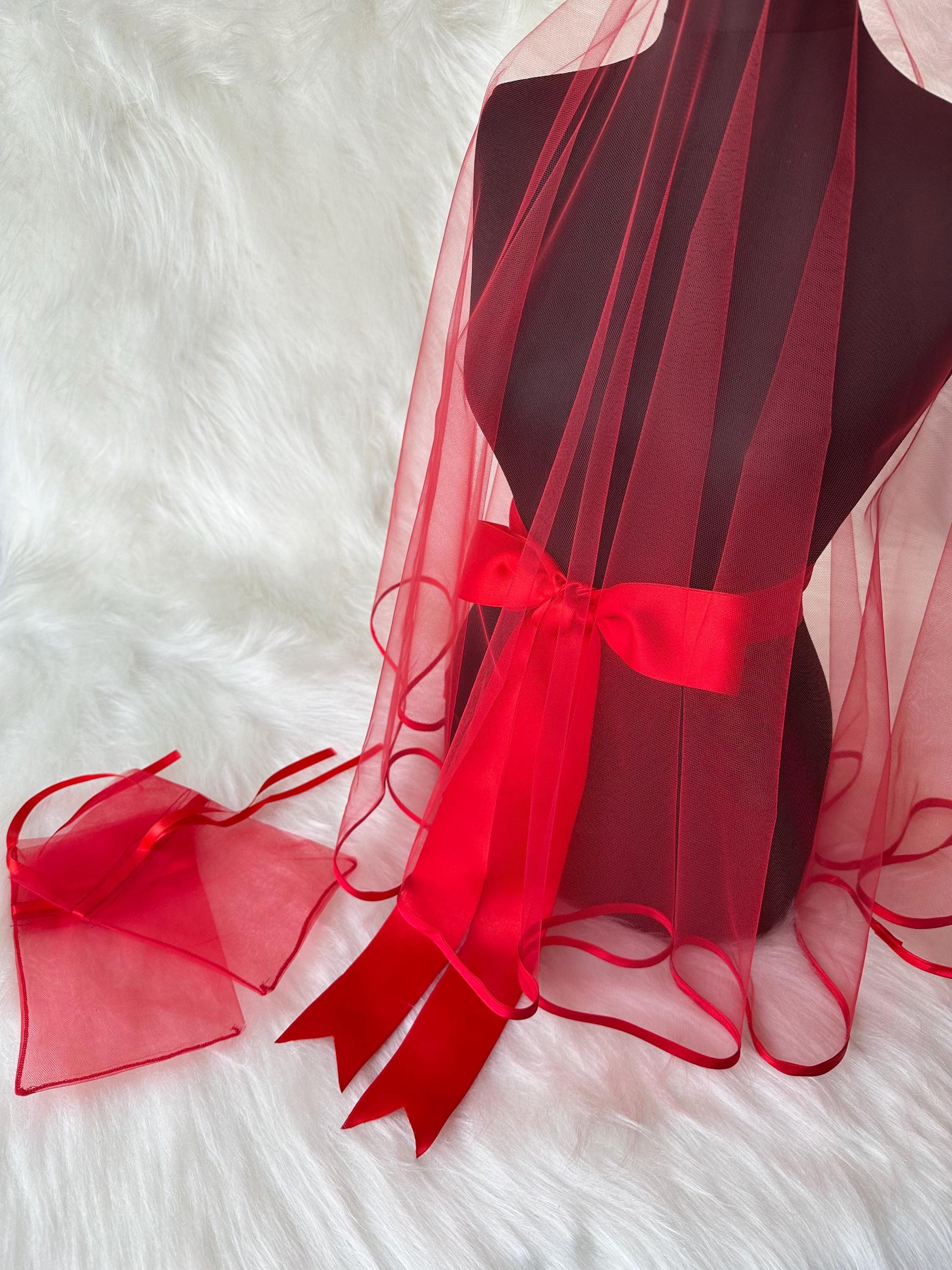 A red tulle veil with a red satin ribbon tied around the center. The veil has a scalloped edge and is displayed on a mannequin. There is also a small red tulle bag with a red satin ribbon.