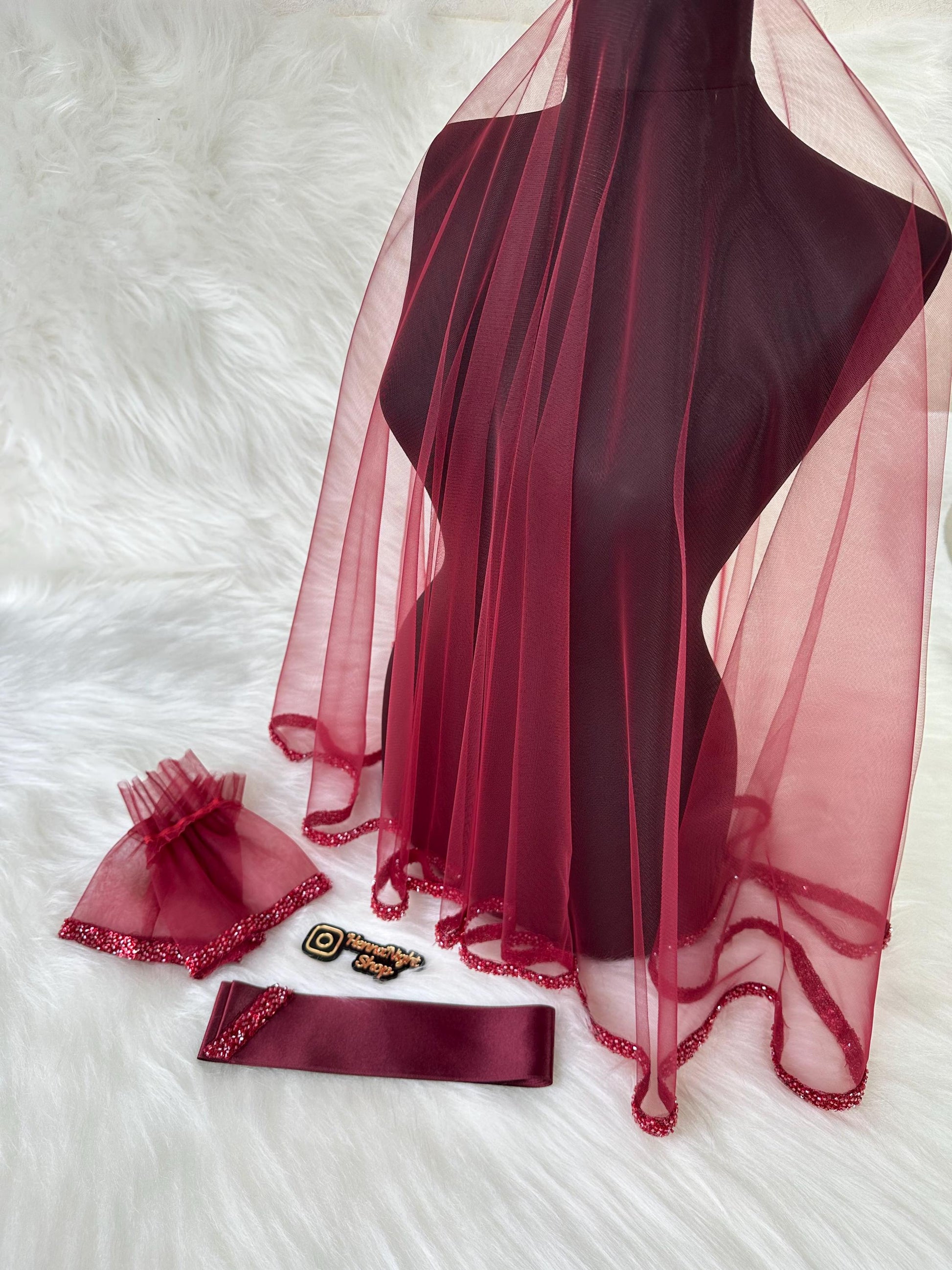 A burgundy bridal veil with a beaded trim. The veil is displayed on a mannequin. There is also a burgundy satin ribbon and a pair of burgundy lace gloves with beaded trim.