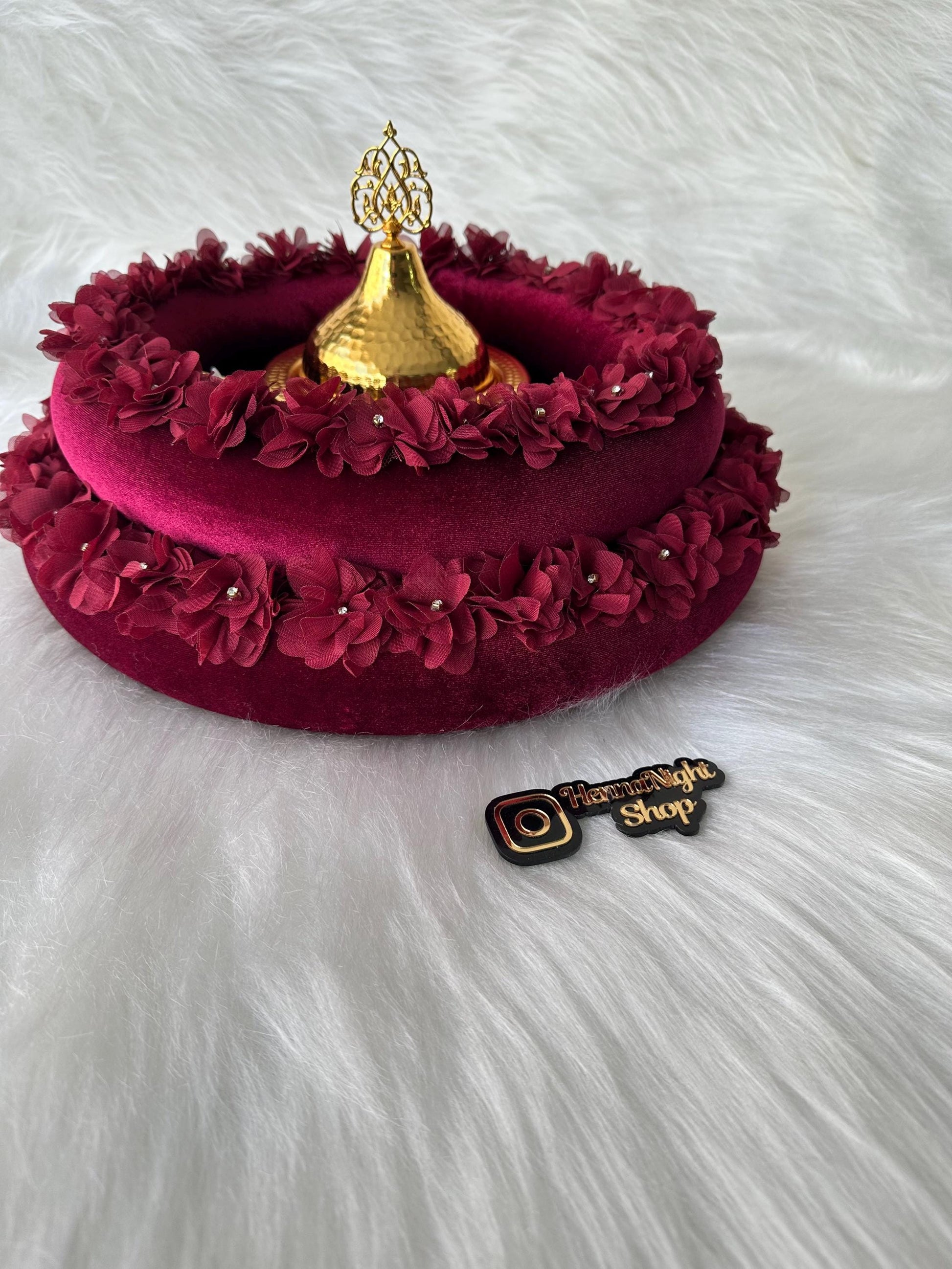 A burgundy velvet tray with a gold metal lid and a decorative floral trim. The tray is decorated with small white flowers. The tray is sitting on a white fluffy surface. The Instagram logo with the words 'HennaNight Shop' is visible in the bottom right corner of the image.