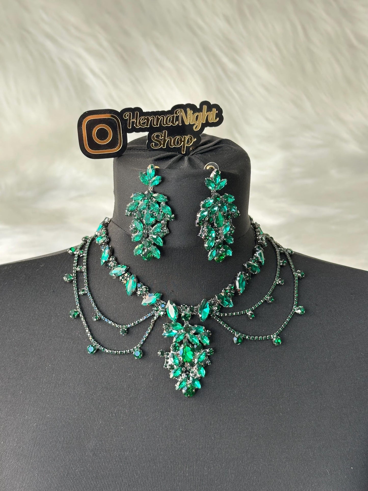 A green forehead jewelry and earrings set with clear and champagne colored rhinestones. The necklace has a layered design with a large pendant in the center. The earrings are dangle style with multiple rhinestones. The jewelry is displayed on a black mannequin bust. The image also includes a logo that says 'HennaNight Shop'.