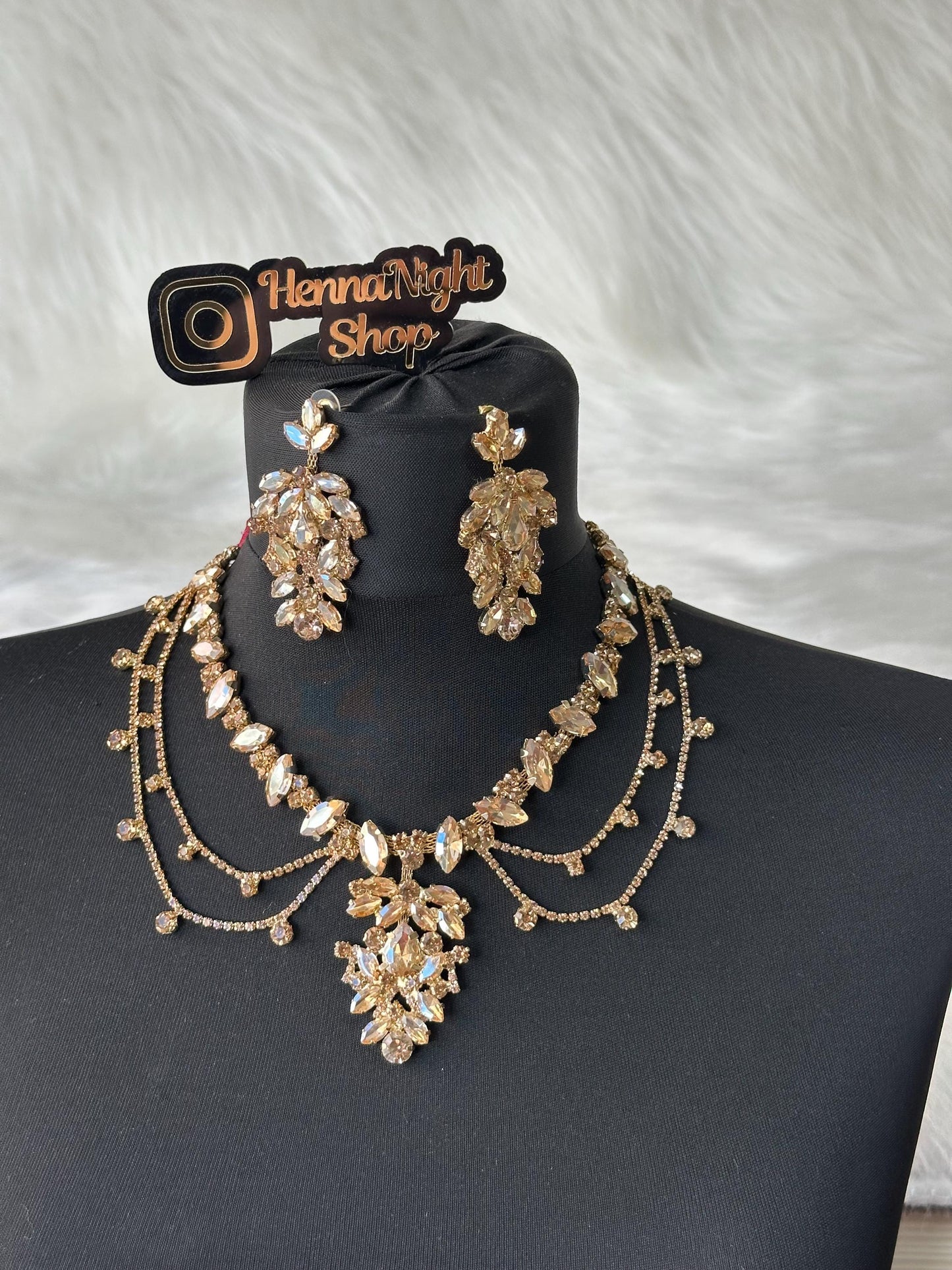 : A gold necklace with a large pendant and matching earrings. The pendant and earrings are made of clear crystals and have a teardrop shape. The necklace has three strands of small crystals. The image includes the text 'Henna Night Shop'.