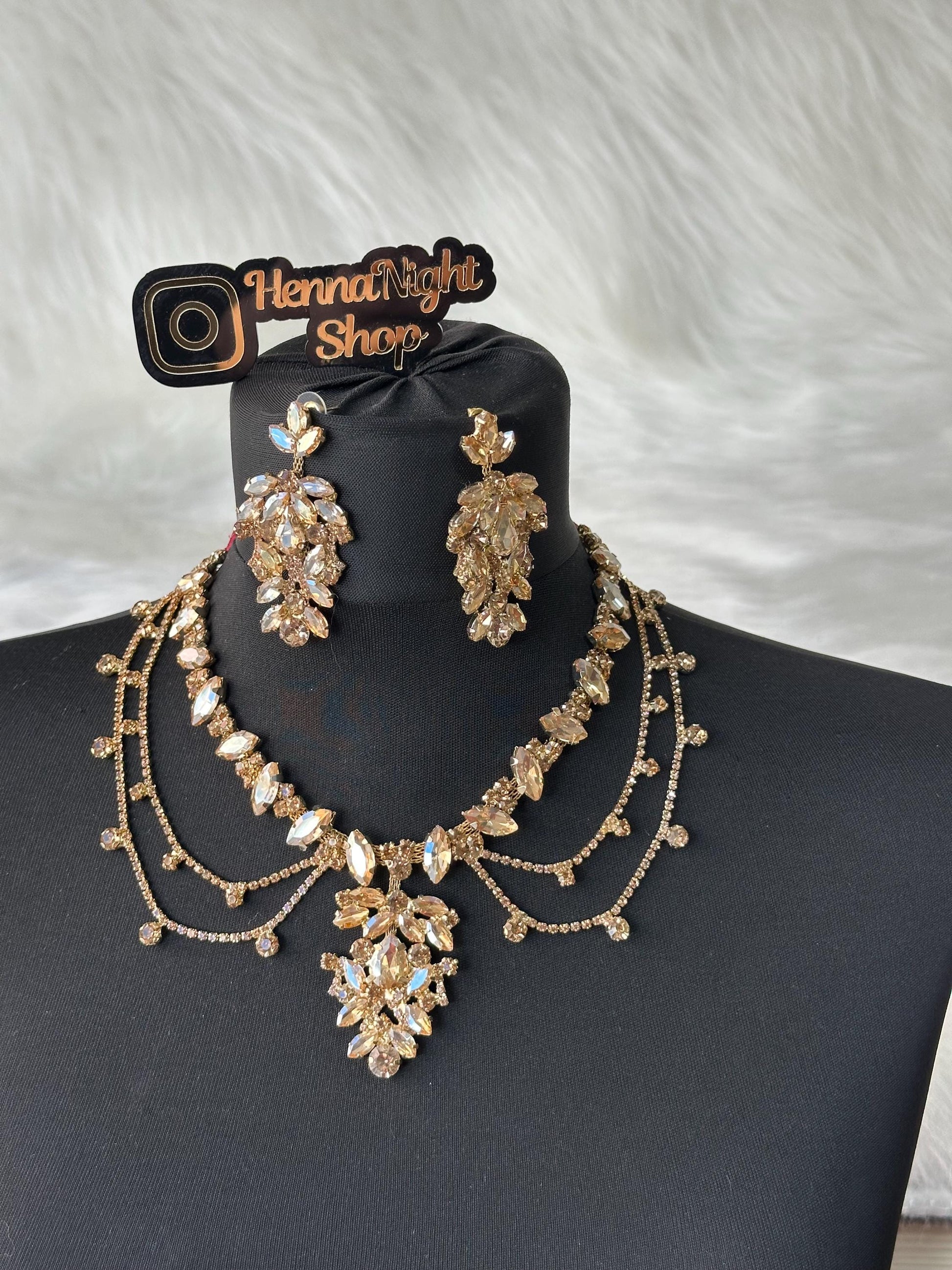 A gold forehead jewelry and earrings set with clear and champagne colored rhinestones. The necklace has a layered design with a large pendant in the center. The earrings are dangle style with multiple rhinestones. The jewelry is displayed on a black mannequin bust. The image also includes a logo that says 'HennaNight Shop'.