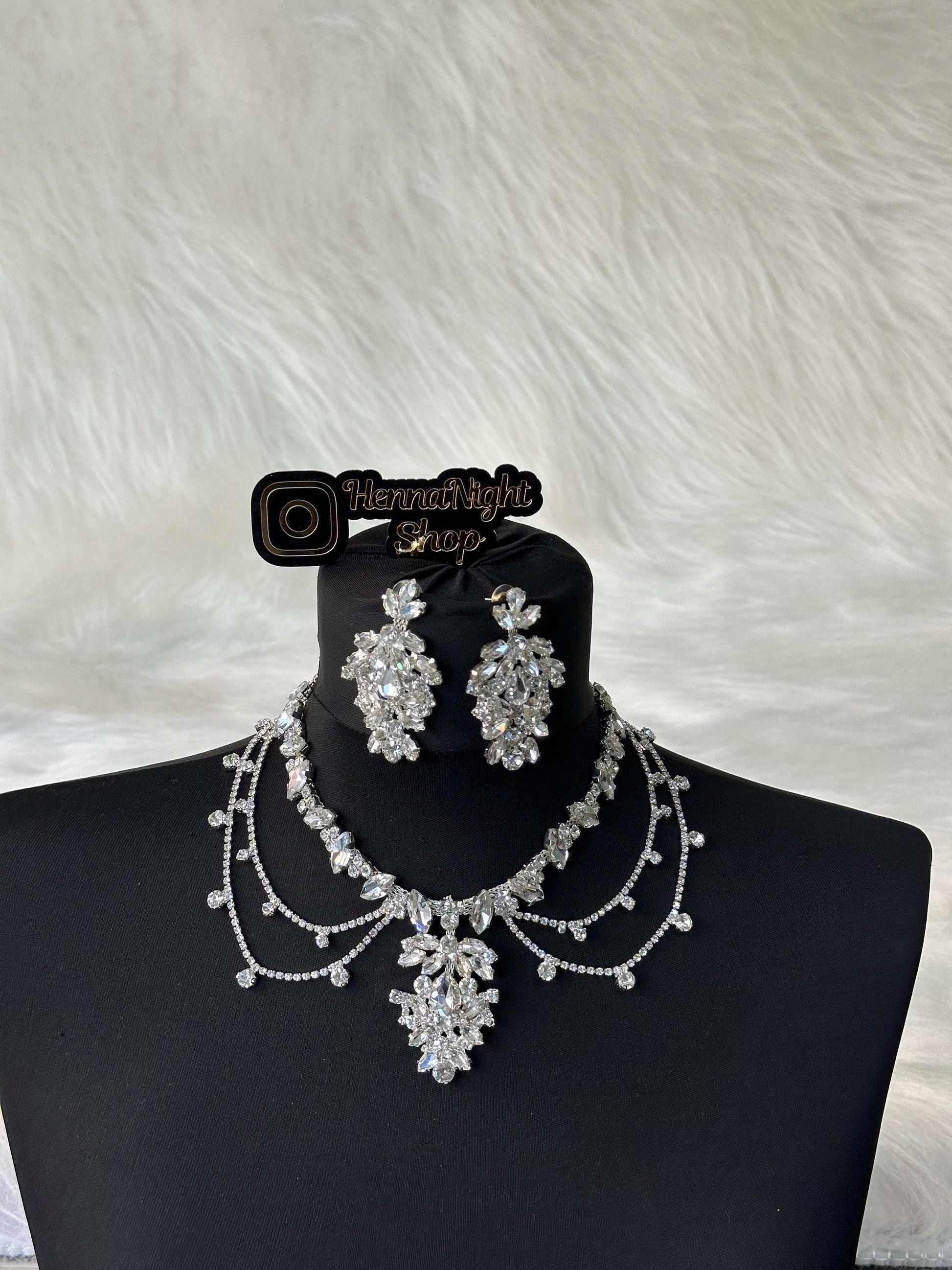 A gold necklace and earrings set with clear and silver-colored rhinestones. The necklace has a layered design with a large pendant in the center. The earrings are long and dangle. The set is displayed on a black mannequin. The text 'HennaNight Shop' is visible in the top center of the image.