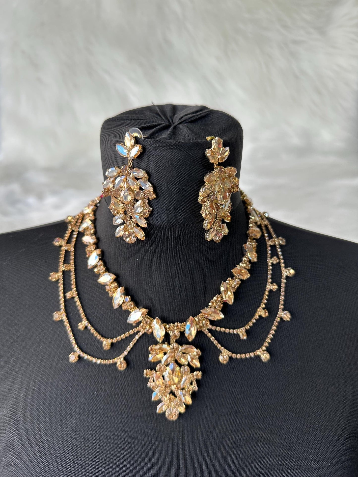A gold necklace and earrings set with clear and golden-colored rhinestones. The necklace has a layered design with a large pendant in the center. The earrings are long and dangle. The set is displayed on a black mannequin. The text 'HennaNight Shop' is visible in the top center of the image.