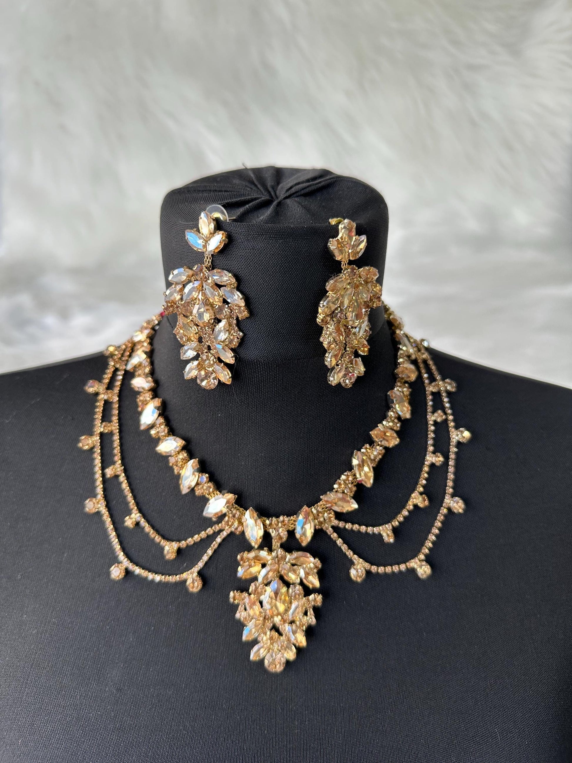 A gold necklace and earrings set with clear and golden-colored rhinestones. The necklace has a layered design with a large pendant in the center. The earrings are long and dangle. The set is displayed on a black mannequin. The text 'HennaNight Shop' is visible in the top center of the image.