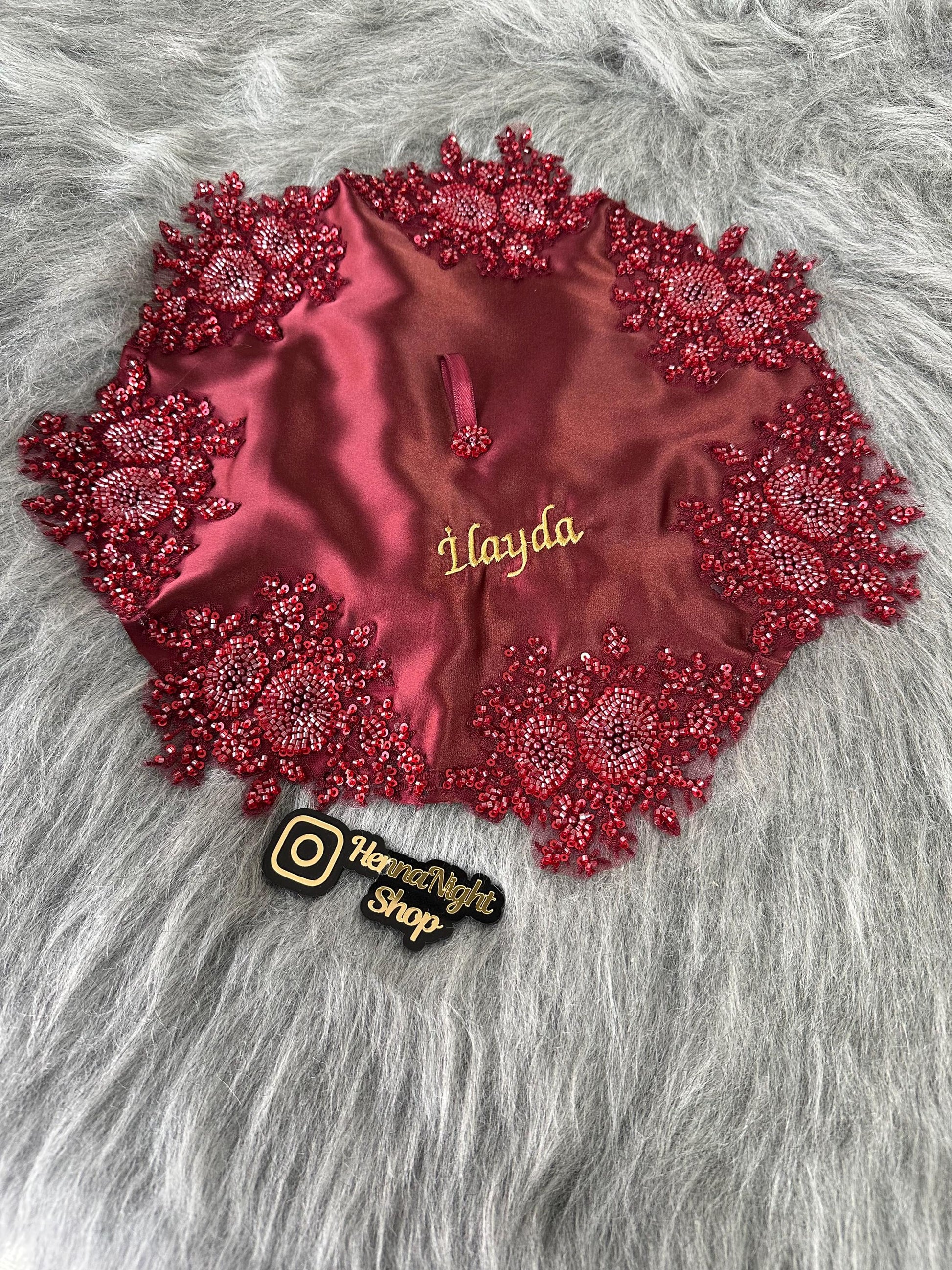 A burgundy satin fabric square with red beaded embellishments around the edges. The fabric square has a gold embroidered name 'İlayda' in the center. The fabric square is on a gray furry surface. A black sticker with a white Instagram logo and the text 'HennaNight Shop' is in the lower right corner of the image.