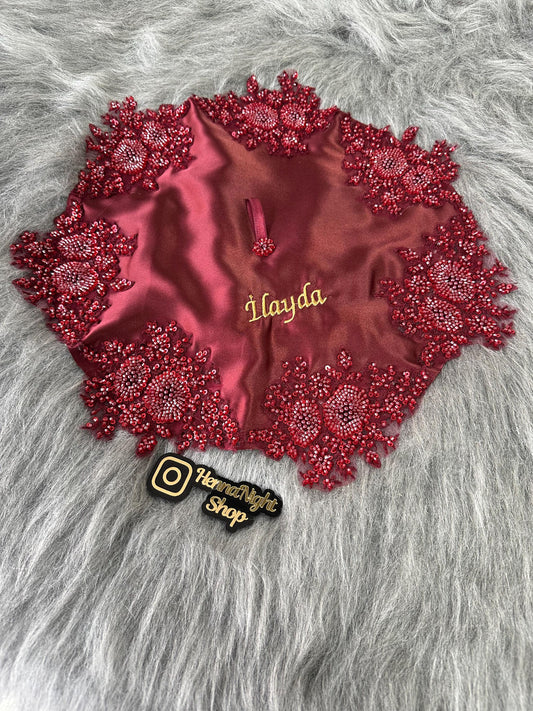 A burgundy satin fabric square with red beaded embellishments around the edges. The fabric square has a gold embroidered name 'İlayda' in the center. The fabric square is on a gray furry surface. A black sticker with a white Instagram logo and the text 'HennaNight Shop' is in the lower right corner of the image.