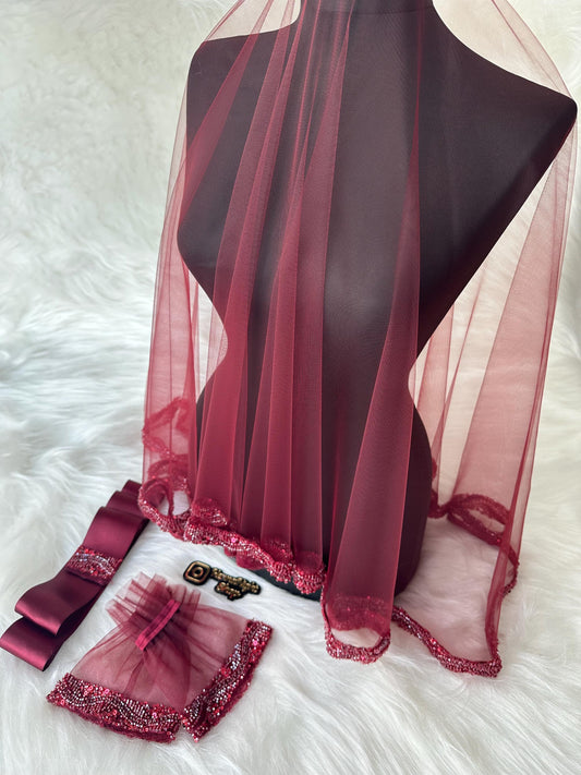 A burgundy tulle veil with a beaded trim. The veil is displayed on a mannequin. There are two matching burgundy tulle wrist cuffs with beaded trim and a burgundy satin ribbon with a beaded trim.