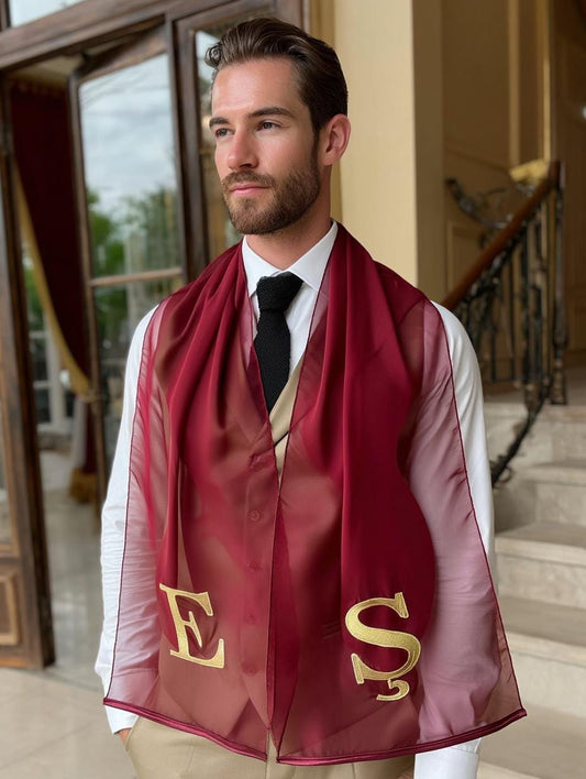 A burgundy scarf with gold embroidered letters "E" and "S" is draped over a white collared shirt and black tie. The scarf is sheer and appears to be made of a lightweight fabric. The person is wearing a white shirt and a black tie