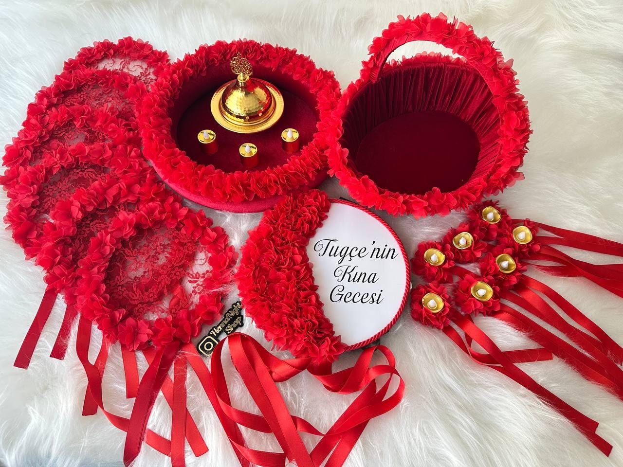 A white round sign with the text 'Tuğçe nin Kına Gecesi' in black lettering. The sign is surrounded by red feathers, red beads, and red fabric. The sign is decorated with a red satin ribbon. The sign is on a white surface. henna tray and henna basket