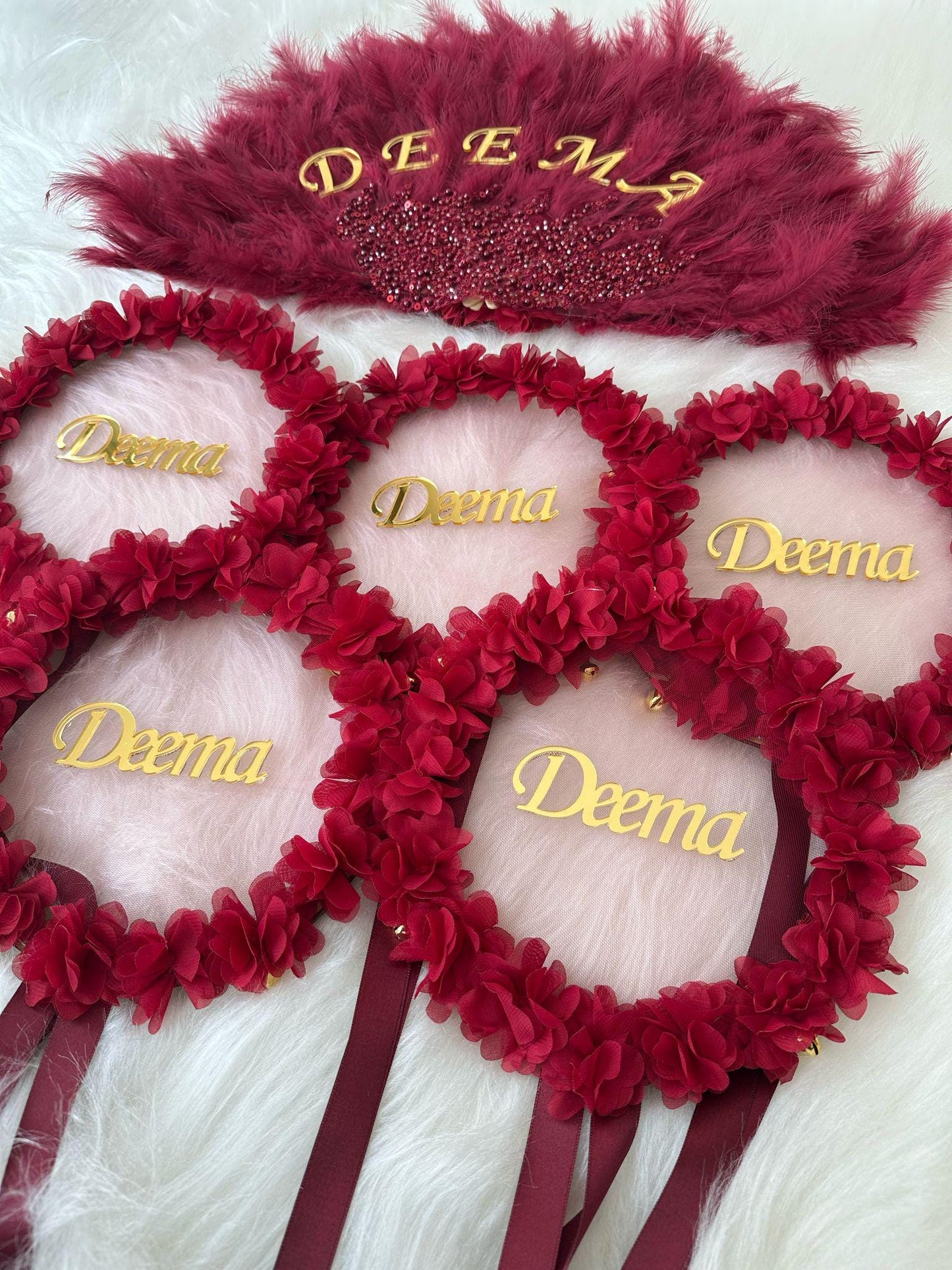 Five red flower wreaths with gold lettering that says 'Deema' on each wreath. The wreaths are tied with burgundy ribbons. A burgundy feather fan with gold lettering that says 'Deema' is in the background.