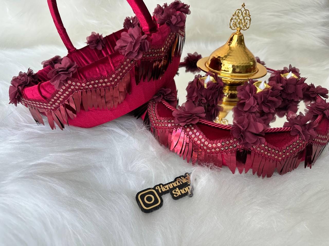 Two decorative trays henna with red velvet and gold accents. The trays are decorated with red velvet fabric, gold sequins, and red fabric flowers. The trays are on a white fluffy surface. The larger tray has a gold bowl with a gold finial on top. The smaller tray is a basket with a handle. The Instagram handle 'Henna Night Shop' is visible on a black tag.