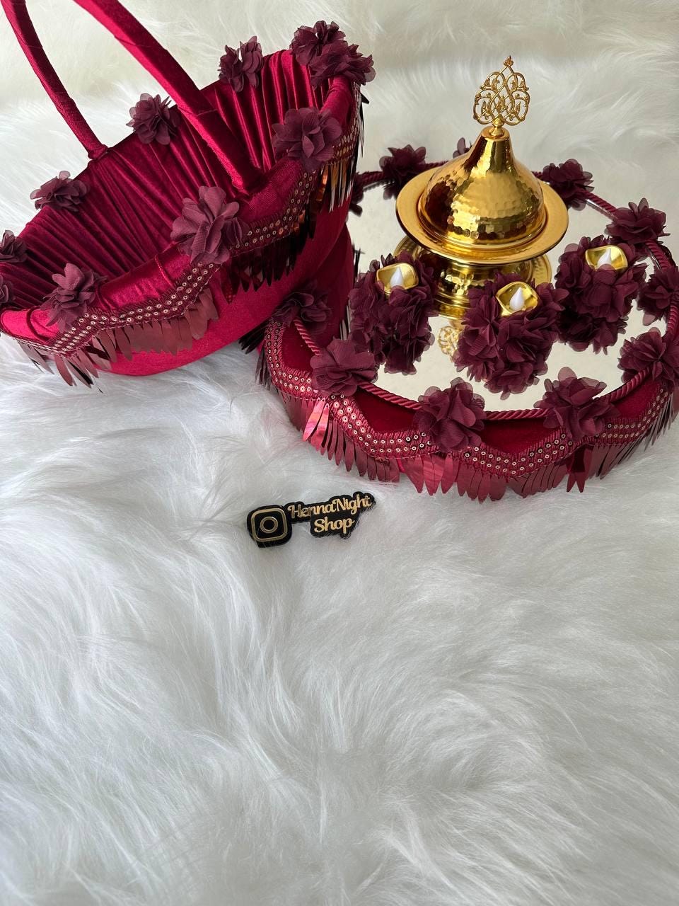 A red velvet henna tray with gold trim and a gold metal bowl with a decorative top. The tray is decorated with red fabric flowers and has a mirror surface. The tray is on a white fluffy surface. The text 'HennaNight Shop' is visible on the surface.