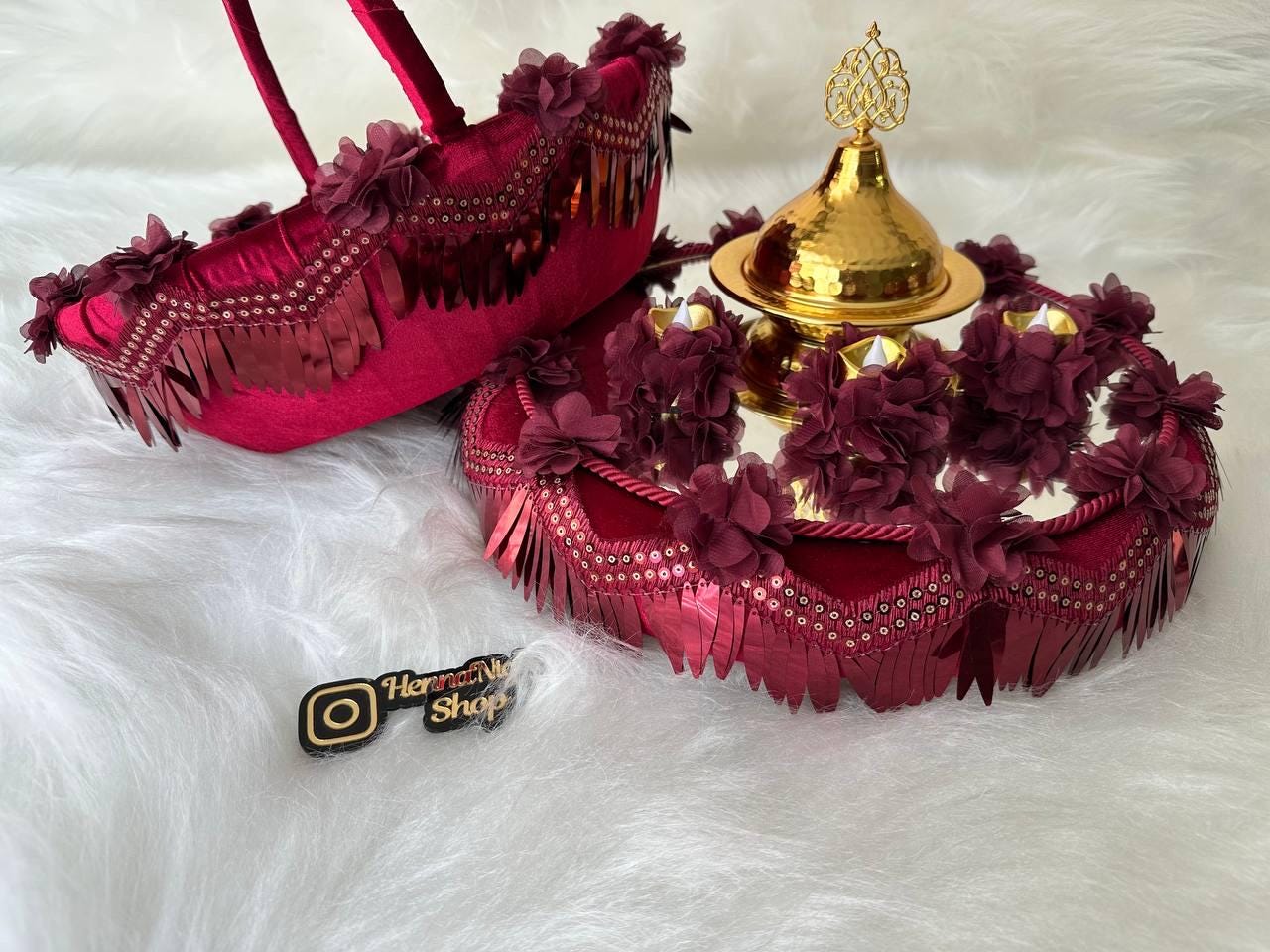 A maroon velvet tray with gold trim and a gold metal bowl with a decorative top. The tray is decorated with maroon fabric flowers and sequins. The bowl is filled with white fabric flowers. The tray is sitting on a white fuzzy surface.