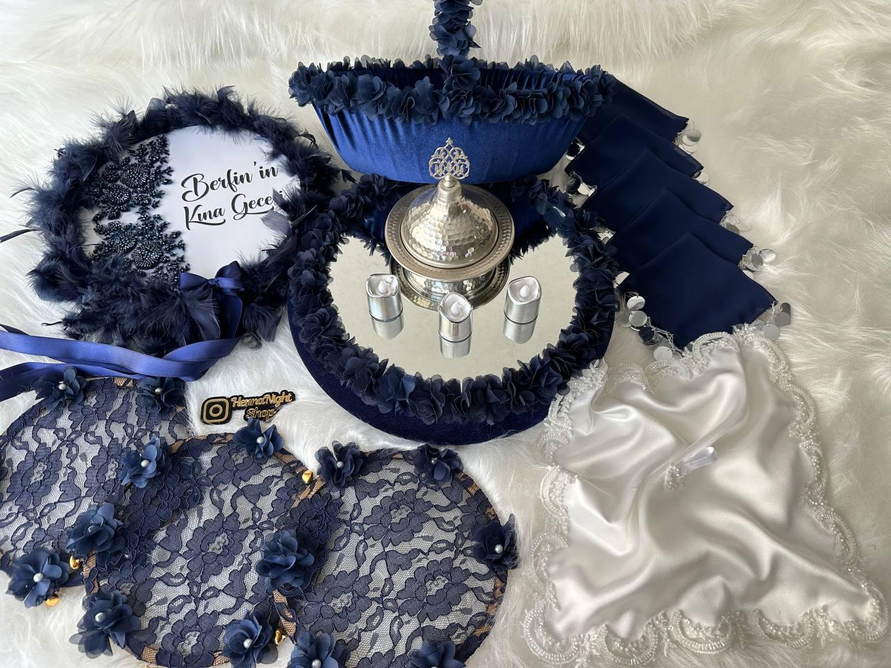 A white round sign with the text 'berfin nin Kına Gecesi' in black  lettering. The sign is surrounded by blue feathers, blue beads, and blue fabric. The sign is decorated with a blue satin ribbon. The sign is on a white surface. henna tray and henna basket blue and white bride handkerchief